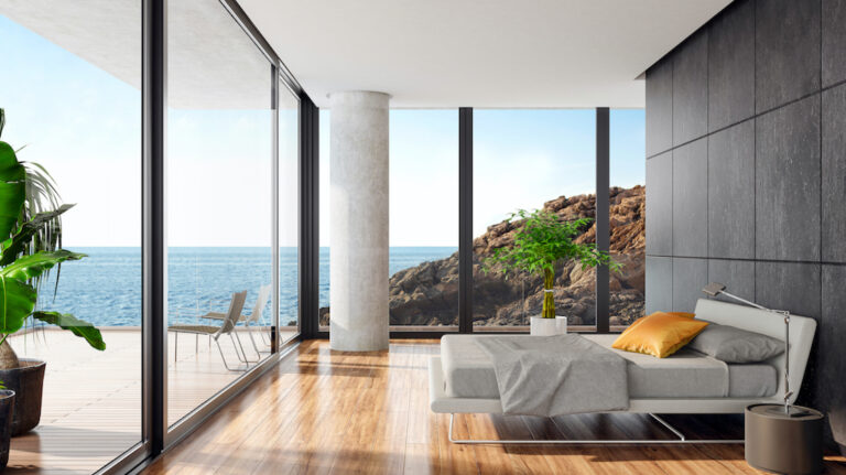 Luxurious minimalist bedroom in a seaside villa with big panoramic windows
and ocean view. White textile king size bed with white metal construction in front of 
black wall with natural stone large tiles. Round modern gray nightstand with a
modern metal lamp next to the bed. Wooden varnished floor. Big terrace with an
ocean view and glass fence with green plants. Summer daylight scene.
Background is my own photo.